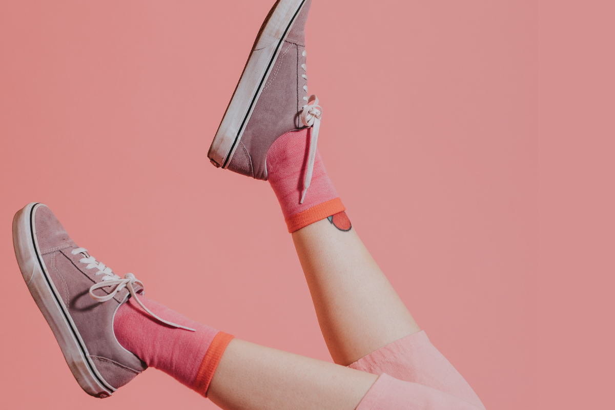 A person wearing light pink pants, bright pink socks with orange trim, and mauve sneakers with white laces is playfully kicking their legs up against a solid pink background.